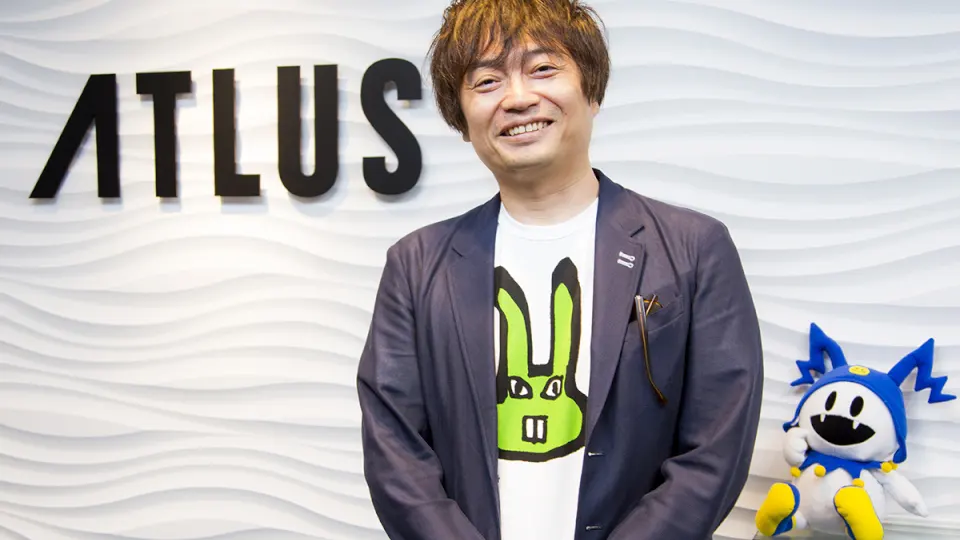 Katsura Hashino sonriente posando delante del logotipo de Atlus